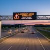 Autoroute estonienne au crépuscule avec un panneau dynamique affichant "ACCIDENT AHEAD" et "EXPECT DELAYS". Des voitures circulent, lumières allumées.